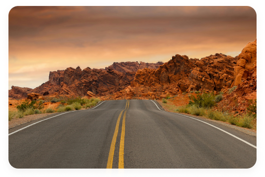 Homepage Feature Image Black tarmac road with yellow stripe down the middle approaching orange-ish hills that look to be in the desert